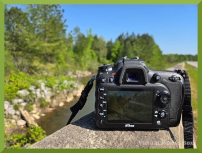 Photo of camera on a wall next to a road, image on camera preview matches creek to left.