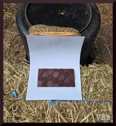 Photo of photography set up. Chocolate bar on a sheet of white paper, on a bale of hay, backed up by an overturned black bucket and a brush.