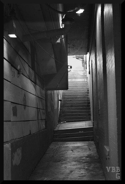 Black and white photograph. Hallway leading to stairs going up into the sunshine.