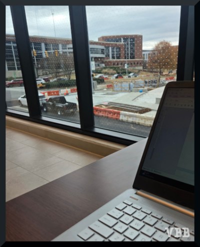 Photo of a window, partial laptop and table in foreground, construction site and buildings in background.