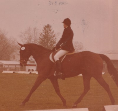 Old photo of rider in black coat and white britches on a bay horse trotting in a grass dressage ring.