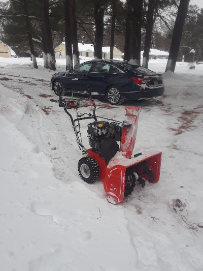 Photo of snow scene with snow blower, cleared driveway, and car, with trees and houses in the background
