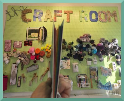 Photo of a notebook held up in front if a wall decorated with craft supplies and titled The Craft Room.
