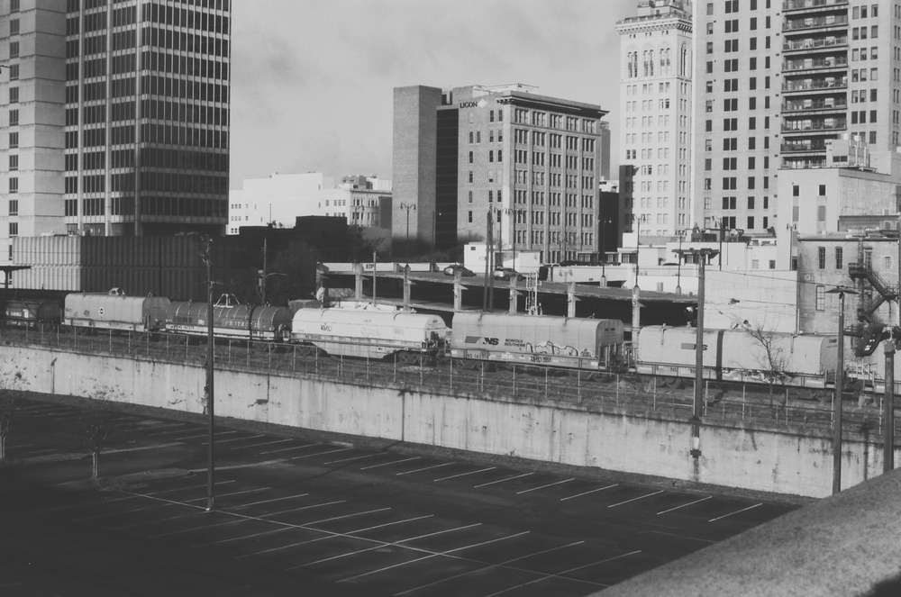 Black and white photo of a downtown area with parking lot, tracks, and buildings.