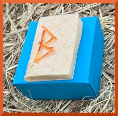 Edible book with the letter B on the cover, resting on a blue paper box resting on a hay bale.