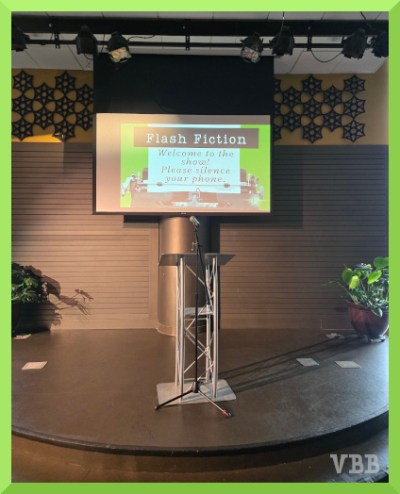 Photo of an empty half-circular stage with microphone, speaker's stand, and plants. In the background, a screen with a slide that says Flash Fiction, Welcome to the show, Please silence your phone.