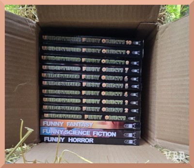 Photo of a stack of 13 paperback books in a open box on its side.