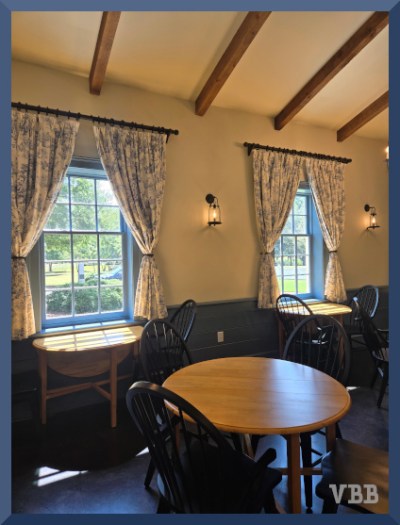 Photo of the interior of Merchants' Coffee House, showing three wooden tables with chairs, two windows with drapes, white walls, blue wainscotting, and wooden beams.