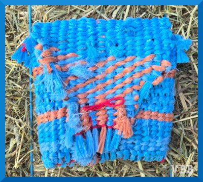 A photo of a woven bag made from blue and orange plastic twine, resting on a hay bale.