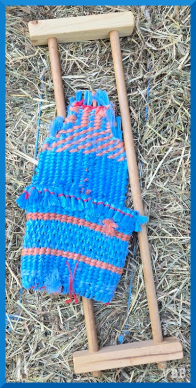A photo of an woven bag made from blue and orange plastic, with the flap open, resting on a wooden loom, resting on a hay bale.