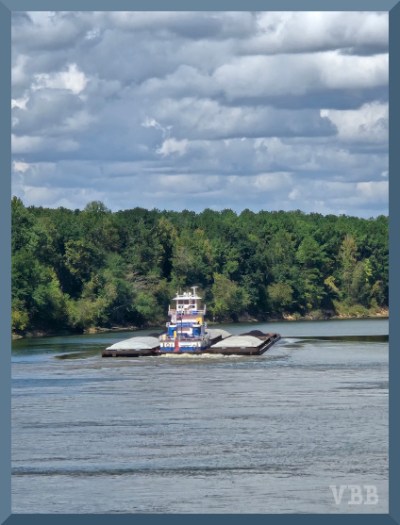 Blurry photo of a tug boat with barges on water with trees in the background and puffy clouds in the sky.