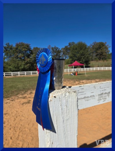 Photo of a blue horse show ribbon and a red horse show ribbon hanging on a monogrammed mug standing on a white fence post, with a red judge's tent and show ring in the background.